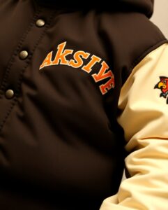 "Close-up of a varsity jacket with 'AKSIVE' embroidered in bold orange and yellow letters on the chest, featuring contrasting cream sleeves and detailed with a small, colorful mascot patch on the sleeve.